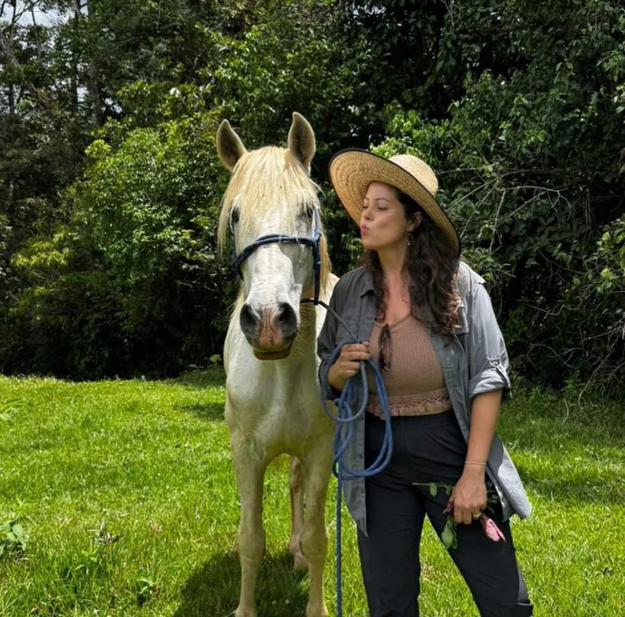 Sara in a sun hat standing with a white horse in a green pasture